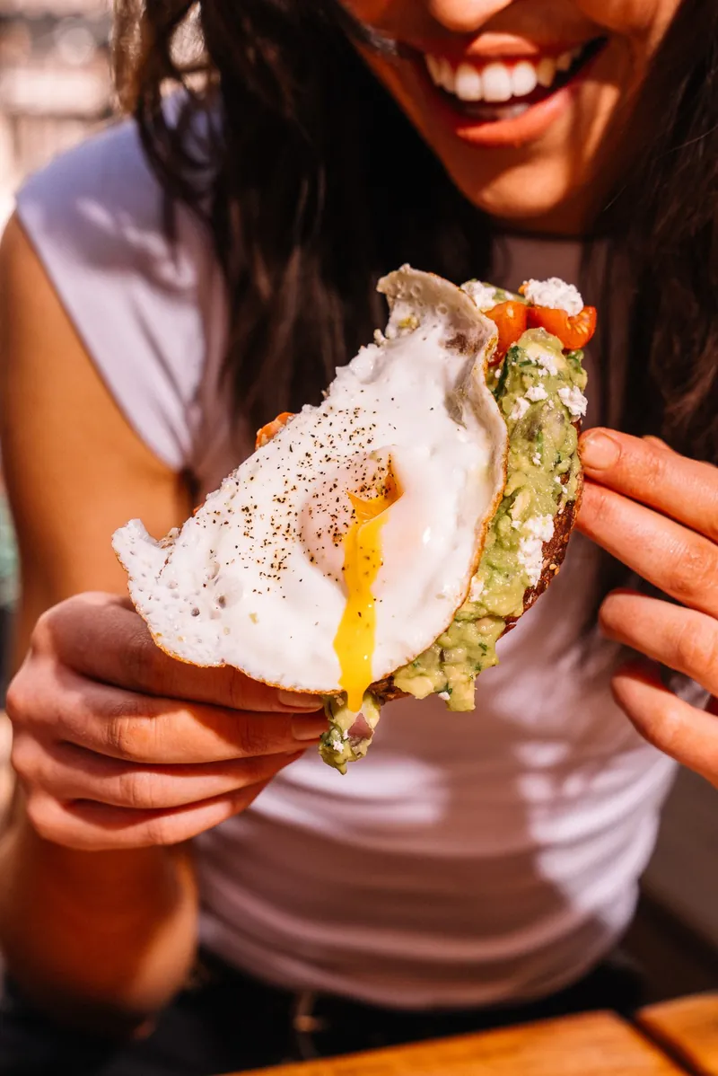 A woman smiling while holding avocado toast with an egg on it