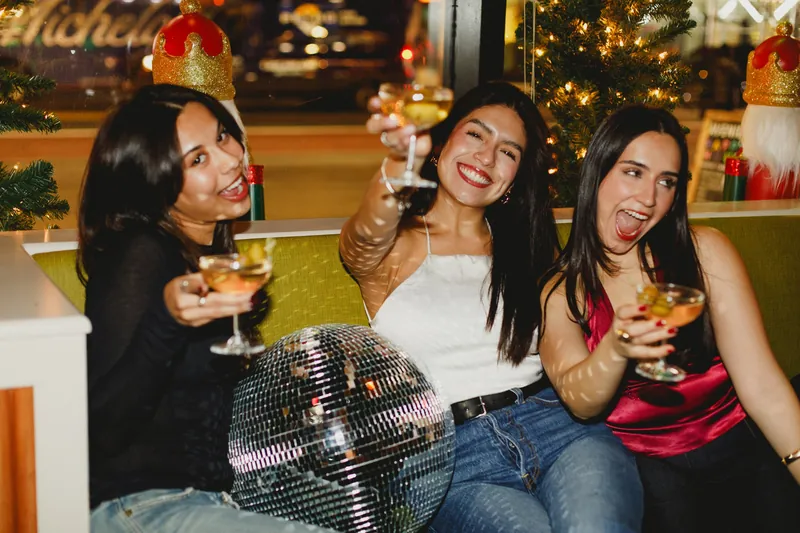 Three women celebrating indoors, all holding martinis towards the camera