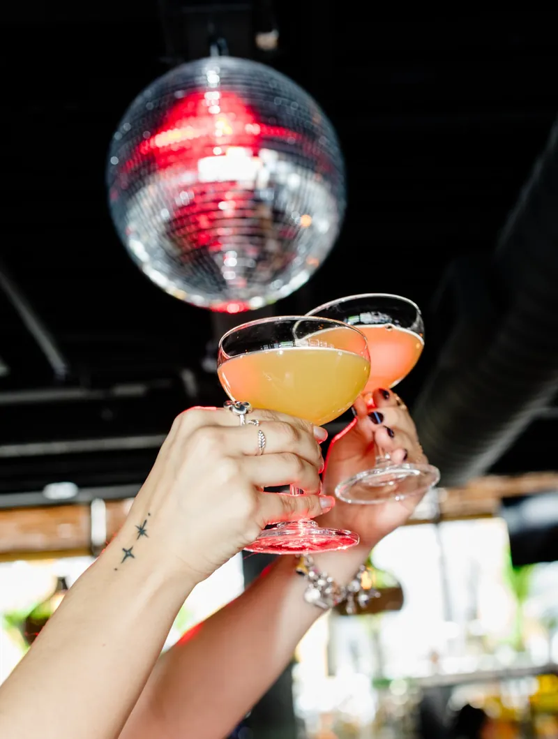 Two cocktails toasting in the air with a disco ball in the background at Avenida